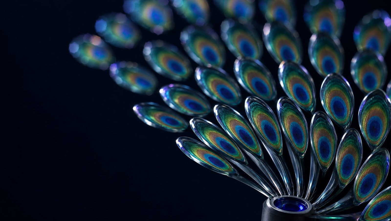 Thirty glass blades set with peacock-feather ocelli fan upward from a small titanium aperture in three arcs; the inner arc is sharp, the outer arc dissolves into soft light.